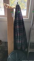 Closed outdoor patio umbrella with striped fabric and plastic weighted base stand in a room near window.