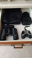 Two pairs of binoculars placed in a drawer with their carrying cases: larger black Bushnell and smaller silver and black Bausch & Lomb.