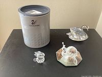 Three Swarovski crystal figurines: a snail on a faceted mirror base in front of a Swarovski branded cylindrical grey case, a swan figurine, and a small bunny figurine.