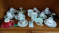 Grouped display of eleven sets of assorted tea cups and saucers, two mugs, and one lone cup on a wooden shelf