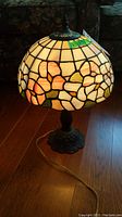 Front view of Tiffany style lamp illuminated on wooden floor showing stained glass floral shade with cream, pink, orange, green hues and metal base.