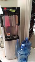 Full view of the Viva self-cleaning water cooler standing on the floor with two blue water bottles next to it.