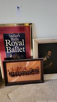 Framed copper 3D artwork of horses and wagon, Royal Ballet poster, and framed portrait painting leaning against a wall.