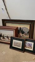 Full view of five framed artworks leaning against a wall, showing a variety of landscape scenes including snowy mountains, trees, and rural settings.