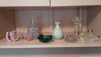 Shelf with multiple glass and ceramic items including carafe with glasses, ceramic figure and animals, glass paperweight.