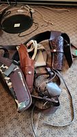 Overall view showing three different gun holsters on a carpeted floor: two leather holsters and one canvas camo holster with straps.