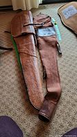 Two long barrel gun cases side by side on carpet floor, one leather carved and one brown fabric with leather accents with measuring tape showing size.