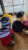 Overview of the assortment of hats showing various types including a black cowboy hat with feathers, a red fez, a furry hat, yellow rain hat, and multiple baseball caps stacked.