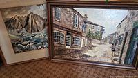 Photo showing both paintings leaning against a wall, framed artwork of a street scene with buildings by Sheila Hersey and mountain scene by Laurie