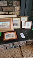 Seven framed art prints arranged on a table against a brick wall, various sizes and frame colors visible.