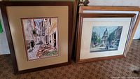 Two framed prints showing street scenes. Left print with a woman walking and crates of produce along a narrow street, right print with a tower in the background and buildings.