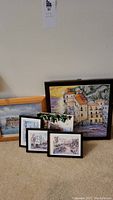 Framed artworks arranged on the floor showing a large framed tile and multiple smaller framed prints featuring village and tropical scenes