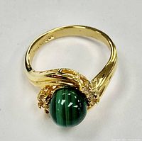 Photo of the front side of the gold electroplated ring featuring a round green malachite gemstone with natural banding held by decorative prongs.