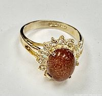 Close up photo of the 18KT gold electroplated ring showing an oval sparkly orange semi-precious gemstone surrounded by clear stones in a floral cluster. Split shank band visible.