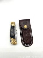 Closed folding knife showing brass engraved bolsters and dark wood handle alongside brown leather sheath with embossed design and brass snap button.
