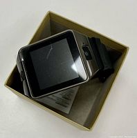 Smartwatch placed inside beige box showing black screen on watch face and black aquatic style silicone band partially visible.