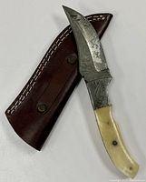 Photo of Damascus steel drop point blade knife with bone handle and dark brown leather sheath with white stitching and rivets.