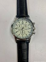 Close-up image of the men's wristwatch showing the blue dial with black numerals, silver bezel with tachymeter, and black crocodile pattern leather band.