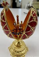 Front view of open egg showing red enameling, gold filigree, and miniature cathedral detail inside on pedestal base.