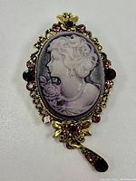 Front view of vintage cameo brooch with a female profile carved in light purple and black, set in an ornate gold-tone frame with pink and red rhinestones and floral design.