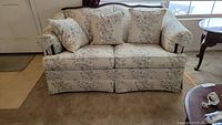 Front facing image of floral cloth loveseat with floral cushions and wood grain accent on backrest, on a beige carpet.