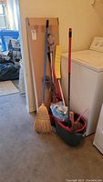 Photo of mop, broom, dustpan, mop bucket with mop heads, and purple telescopic window cleaner leaning against a washer
