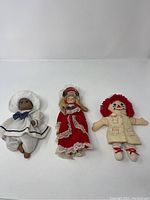 Three dolls including a black sailor doll, European style doll in red dress, and Raggedy Ann doll.