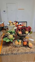 Wide shot of entire assortment of faux fruit in various decorative containers including silver-tone bowls, glass bowl, and loose fruit on countertop.