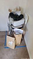 Photo of collapsible walker with padded seat, white toilet seat raiser, foot brace, cushion, and cardboard boxes stacked in a corner.