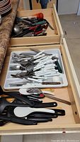 Two drawers showing assorted kitchen utensils and flatware including utensils with white plastic handles and black plastic cooking tools