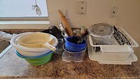 Overview of kitchen items on countertop including white dish drainer, plastic bowls, colander, rolling pin, blue measuring cups and spoons, and wire basket.
