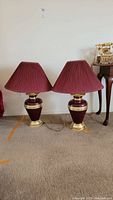 Pair of two red tone ginger jar style lamps with brass type bases and matching pleated red lampshades on a beige carpet.