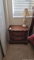 Nightstand with wood grain finish and brass tone drawer pulls positioned next to a window.