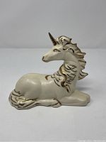 Side and angled views of a reclining porcelain unicorn figurine with cream-white and gold detailing, showing flowing mane and tail