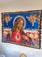 Full view of the religious tapestry hanging on a wall showing Jesus with a glowing sacred heart, angels, clouds, and Middle-Eastern buildings in the background with a decorative border.
