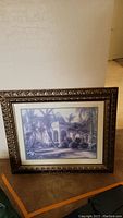 Framed soft black and blue toned print of tropical home under palm trees, with gold tone wood grain textured frame.