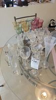 Assorted clear and colored stemware on a glass table including wine glasses, champagne flutes, and shot glasses.