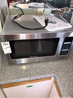 Full view of Moulinex microwave oven on countertop with power cord and paper manual on top, showing stainless steel finish and black door with logo.