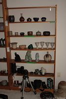 Full view of the tall wooden shelving unit against a wall, showing five wooden shelves holding various small decorative items and photography equipment on the bottom shelf.
