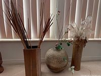 Three decorative ceramic vases placed on a carpeted floor in front of vertical blinds. One rectangular, one large round, one tall cylindrical vase, each with dried plants inside.