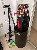 Full view of walking sticks, umbrellas, and black cylindrical stand with some boxed items on the floor.