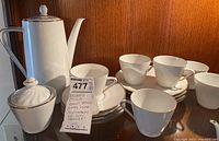 View of the entire tea set including teapot, sugar bowl with lid, multiple teacups with saucers and plates, displaying the white porcelain with ribbed design and gold trim.