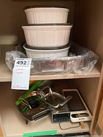 Three white ceramic casseroles stacked, rectangular roaster with rack wrapped in plastic underneath, and assorted graters in front
