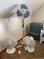 Wide shot showing three fans: one tall standing Glacier brand fan with blue blades and two smaller white desk fans, placed on carpeted floor near wall.