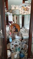 Glass cabinet shelf containing crystal mini vases, collector plates, glass candle holders, and small figurines