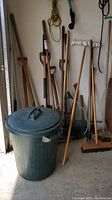 Garden tools arranged against a wall in a garage including garbage bin and long handled tools like shovels and brooms