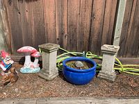 Green garden hose coiled on ground behind two weathered concrete pedestal stands, blue ceramic plant pot with soil and small plants, and three garden ornaments (gnome, mushrooms, squirrel) arranged in front of wooden fence.