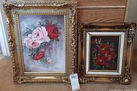 View of two framed floral oil paintings side by side on floor showing overall condition and framing.