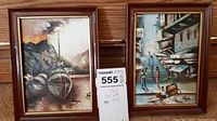 Two framed oil paintings resting on wooden surface: left shows boats on water, right shows street scene with people. Both have brown wooden frames.