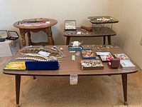 Full shot of mid-century modern coffee table in front with wood top surfaces and metal supports on the upper tier, surrounded by various jewelry. Wooden side table with turned legs seen to left.